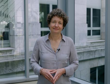 A woman with short curly hair in a grey button down shirt speaks to the camera